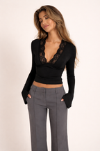 Vrouw draagt zwarte aansluitende top met diepe kanten V-hals en lange mouwen gecombineerd met een elegante grijze pantalon