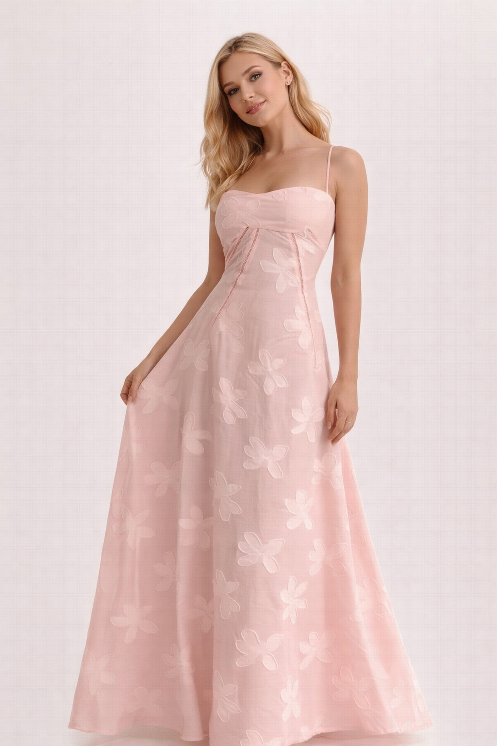 Lichtroze maxi jurk met bloemstructuur en fijne schouderbandjes tegen een witte muur